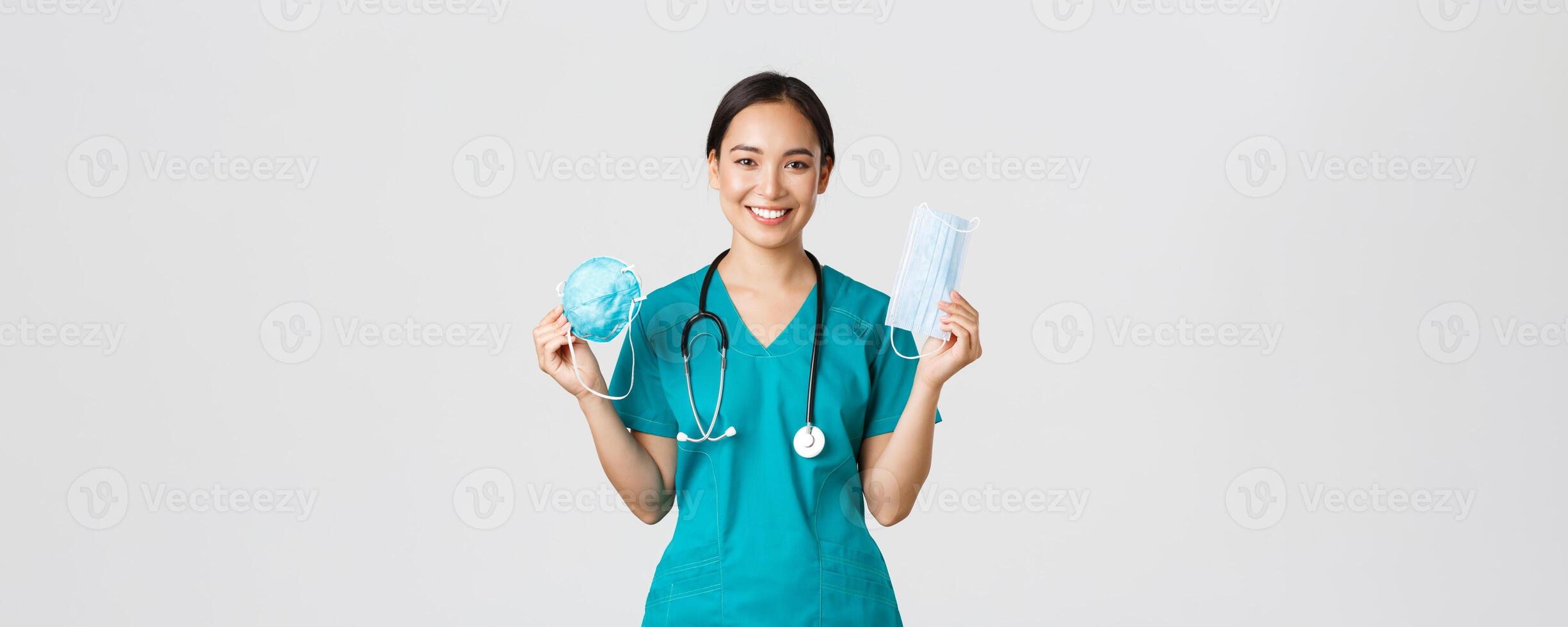 Covid-19, coronavirus disease, healthcare workers concept. Smiling pretty asian female nurse, physician in scrubs showing respirator and medical mask, suggest patients ways of virus protection photo