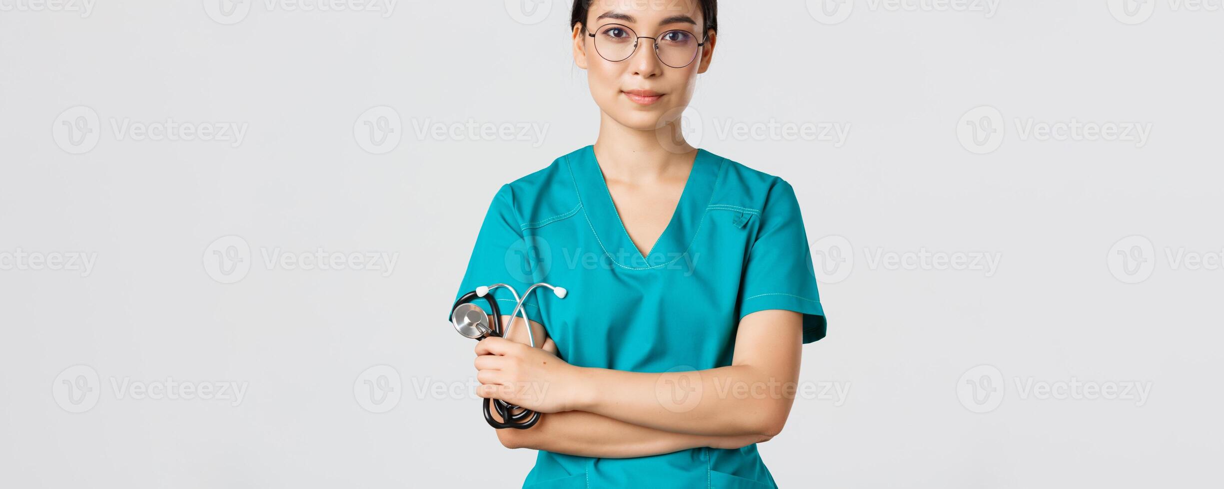 COVID-19, coronavirus enfermedad, cuidado de la salud trabajadores concepto. confidente sonriente profesional asiático médico en anteojos, cruzar brazos pecho, vistiendo matorrales y participación estetoscopio, blanco antecedentes foto