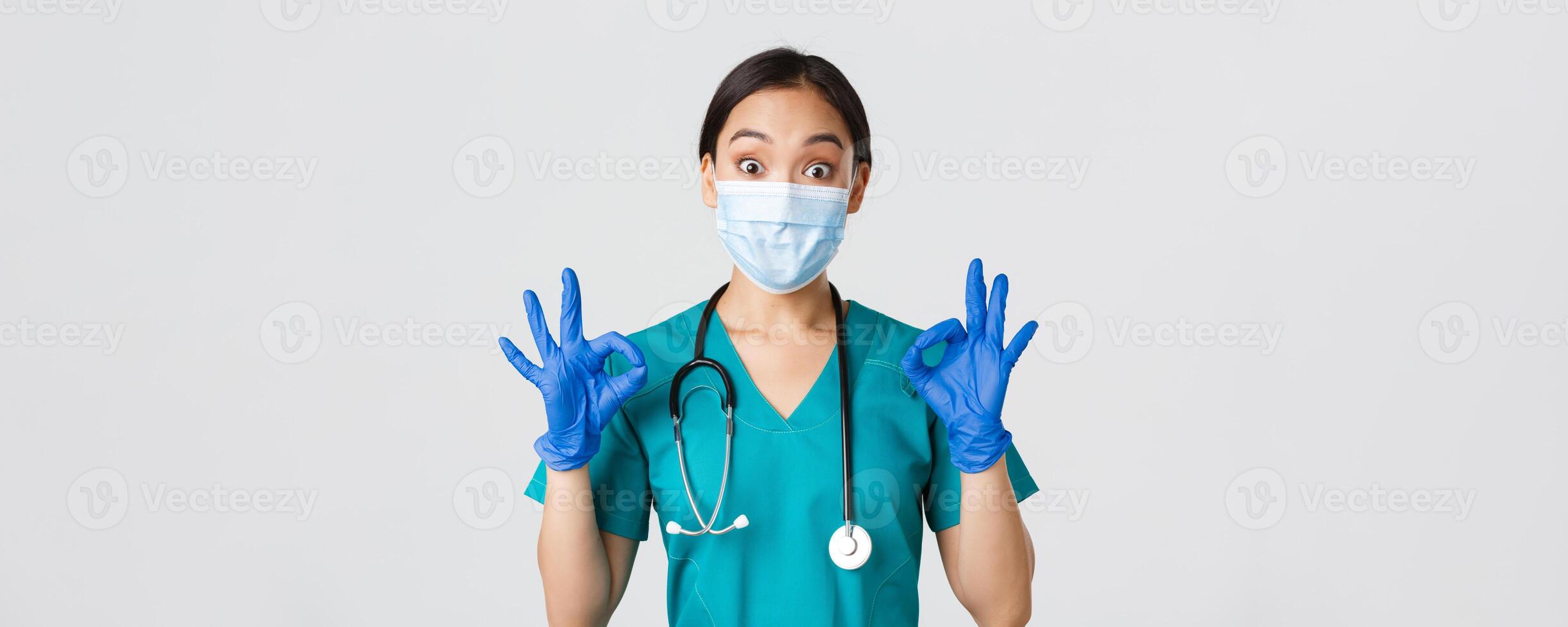 Covid-19, coronavirus disease, healthcare workers concept. Excited and impressed asian doctor, nurse in medical mask and rubber gloves showing okay gesture, approve or praise nice work photo