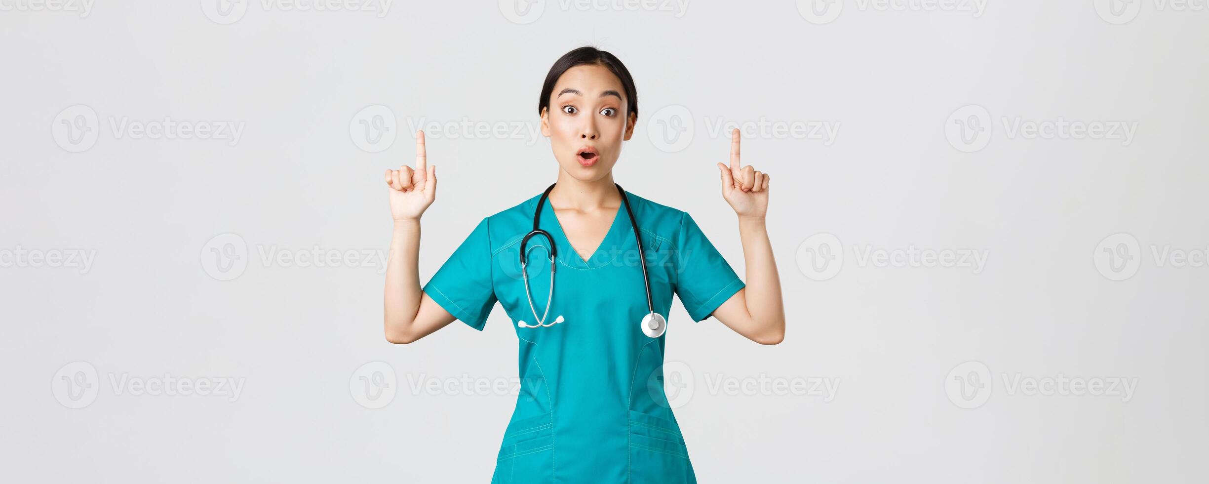 Covid-19, healthcare workers, pandemic concept. Surprised and thrilled asian female nurse, woman doctor in scrubs asking question interesting promo, pointing fingers up, showing clinic advertisement photo