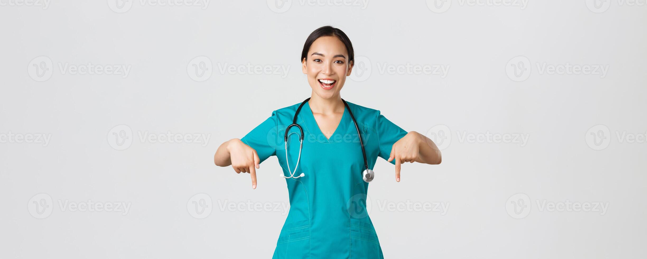 Covid-19, healthcare workers, pandemic concept. Happy smiling beautiful asian nurse, female doctor in scrubs pointing down, inviting for checkup, showing hospital advertisement, white background photo