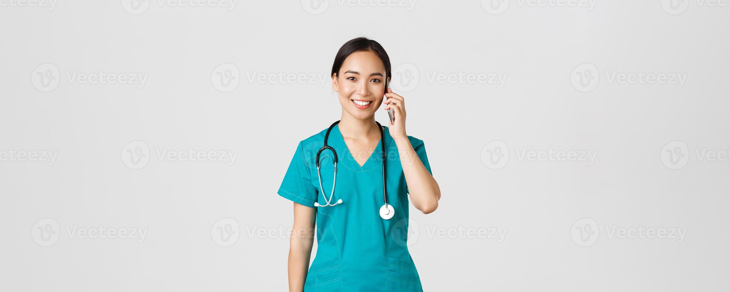 Covid-19, healthcare workers and preventing virus concept. Smiling happy asian female doctor, young intern in scrubs talking on phone, looking at camera hopeful, white background photo