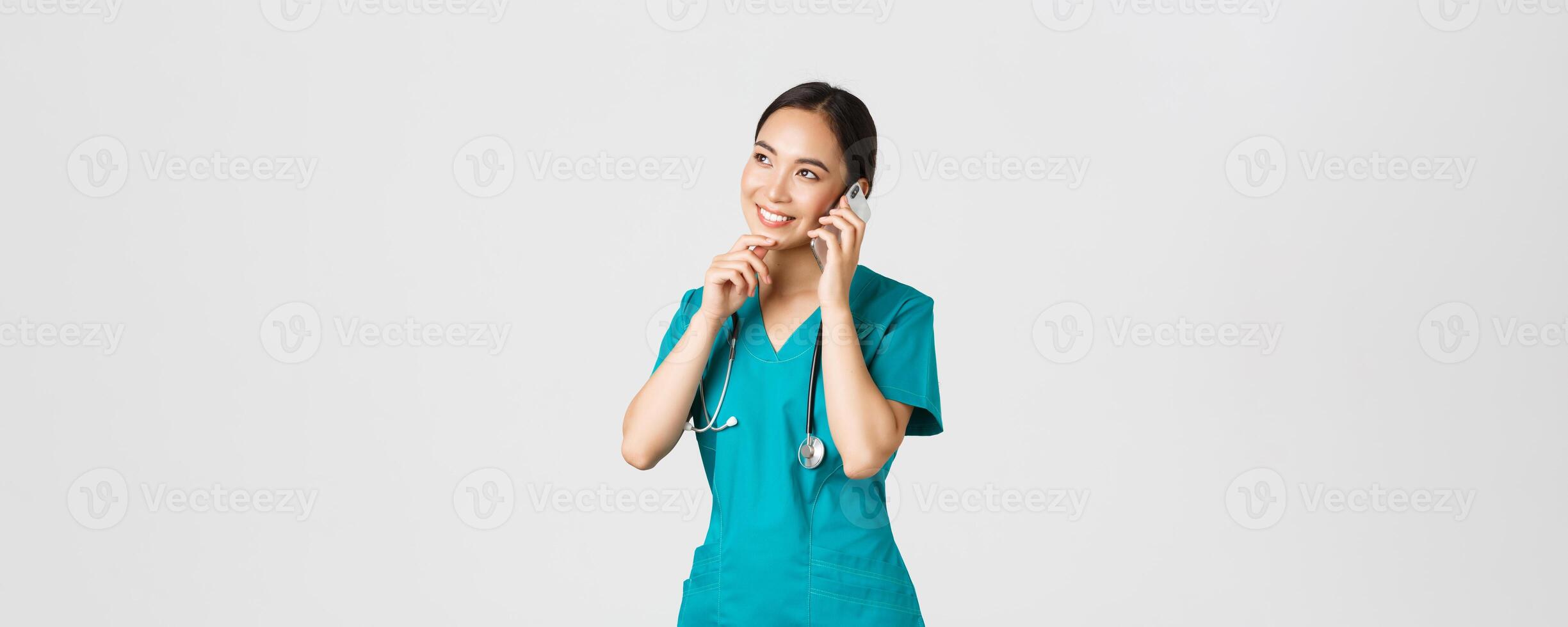 Covid-19, healthcare workers and preventing virus concept. Portrait of smiling asian female doctor, intern in scrubs talking on phone and looking thoughtful, thinking or making choice photo