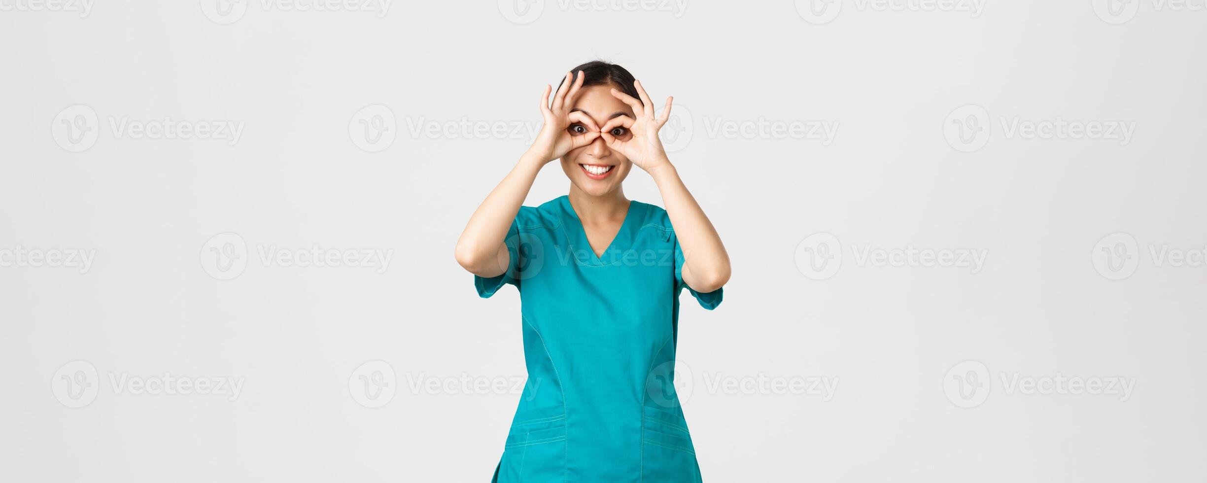 Covid-19, healthcare workers and preventing virus concept. Amused happy, cute asian female doctor, intern in scrubs looking through hand binoculars with excited expression, smiling photo