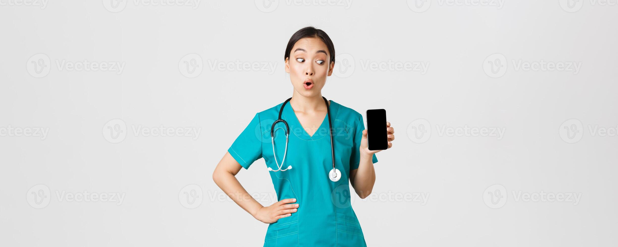 COVID-19, cuidado de la salud trabajadores y en línea medicina concepto. emocionado y asombrado asiático hembra enfermero, médico Mira sorprendido mientras demostración móvil teléfono pantalla, Internet consulta aplicación, blanco antecedentes foto