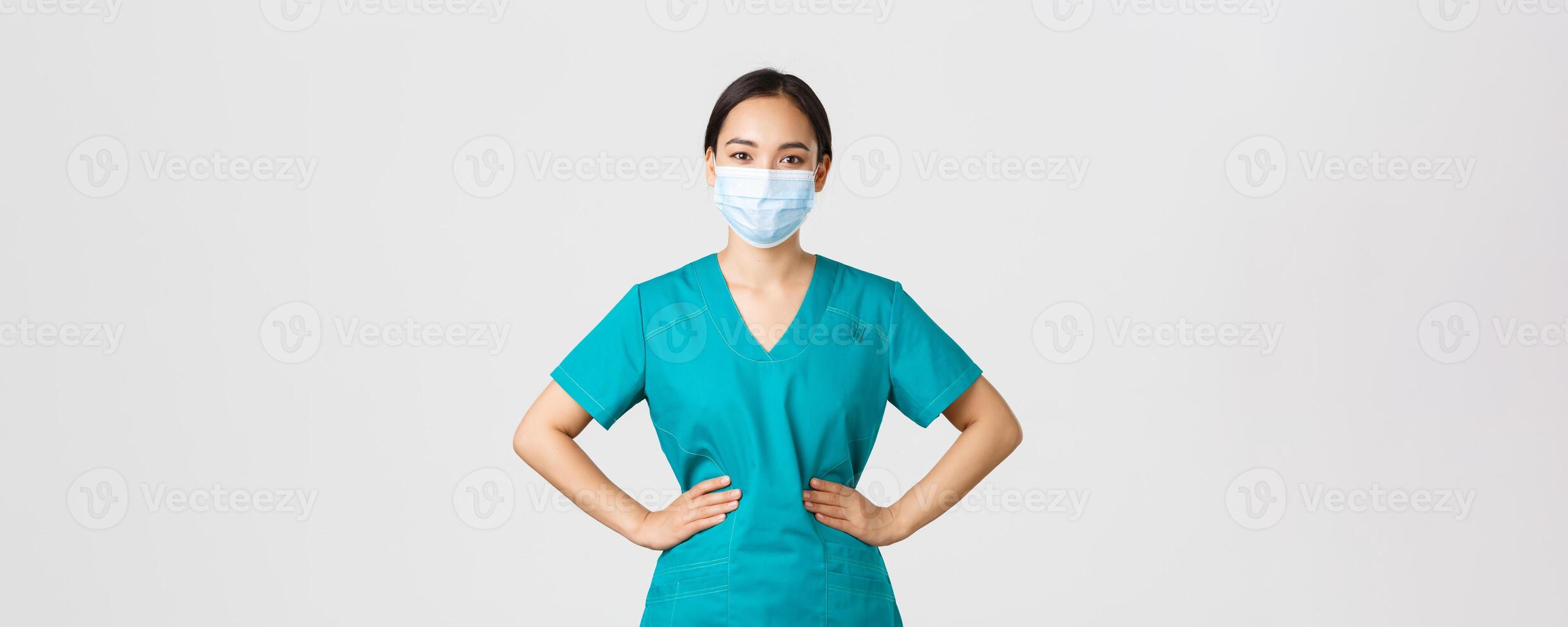 Covid-19, coronavirus disease, healthcare workers concept. Cheerful smiling asian female doctor, physician in scrubs and medical mask, looking upbeat, working with patients in hospital photo