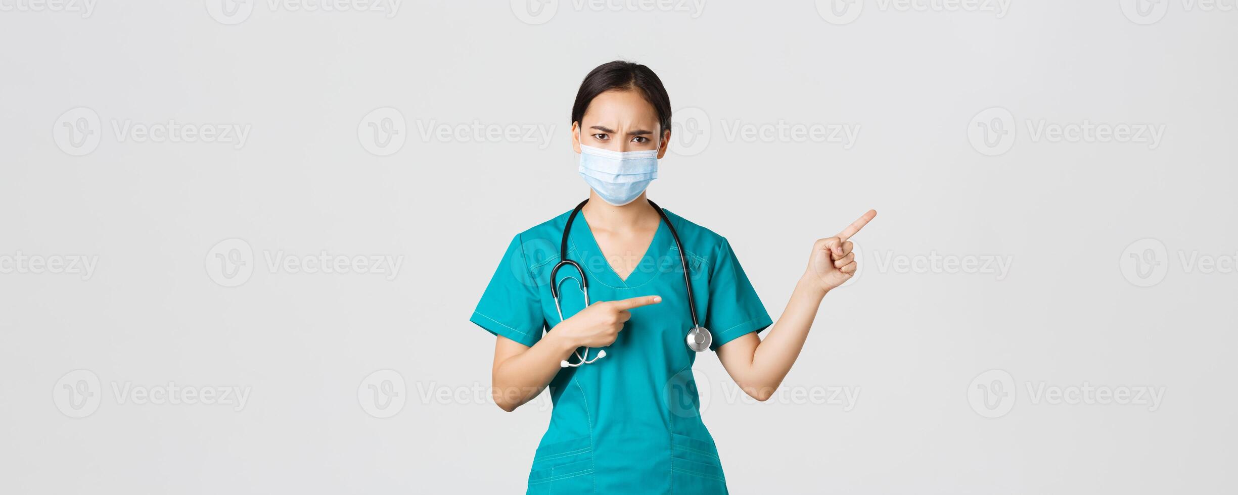 Covid-19, coronavirus disease, healthcare workers concept. Frowning suspicious or frustrated asian female doctor, wear medical mask and scrubs, pointing upper right corner, white background photo