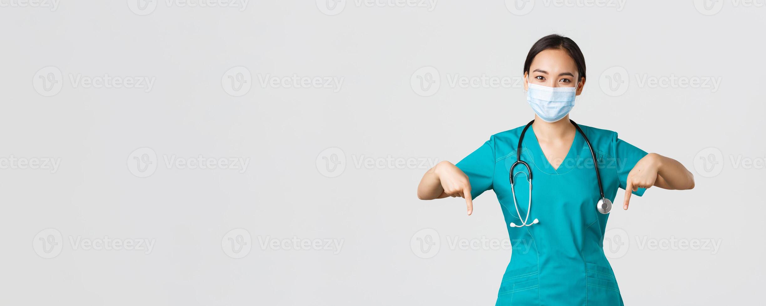 Covid-19, coronavirus disease, healthcare workers concept. Pleasant smiling asian female nurse, doctor in medical mask and scrubs, pointing fingers down, provide information, invite for checkup photo