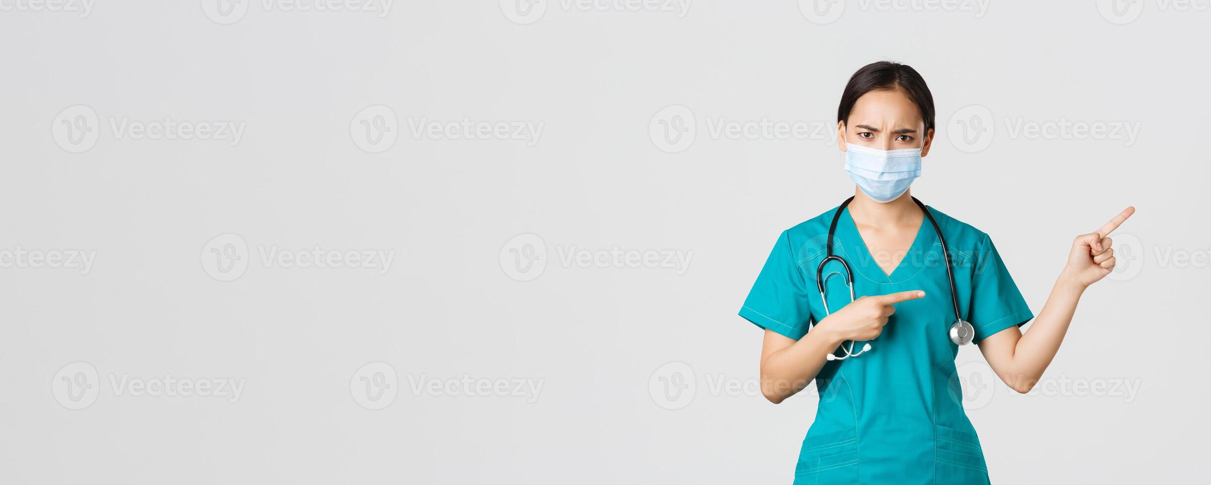 Covid-19, coronavirus disease, healthcare workers concept. Frowning suspicious or frustrated asian female doctor, wear medical mask and scrubs, pointing upper right corner, white background photo