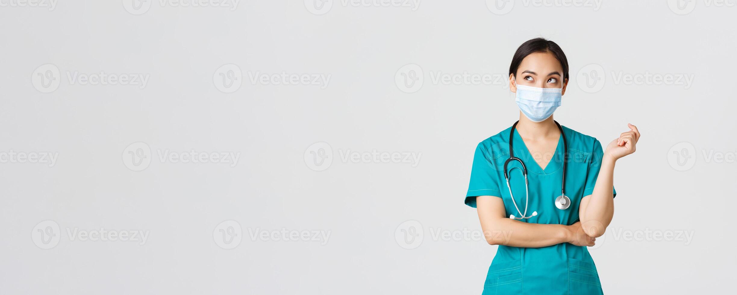 Covid-19, coronavirus disease, healthcare workers concept. Curious and thoughtful asian female doctor in medical mask and scrubs, looking upper left corner, thinking, white background photo