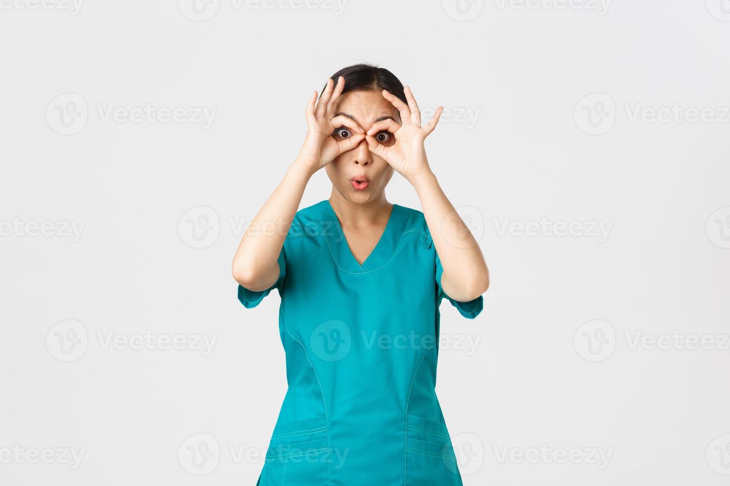 Covid-19, healthcare workers and preventing virus concept. Impressed funny and cute female asian nurse in scrubs looking through fingers over eyes as if stare in binoculats amazed photo