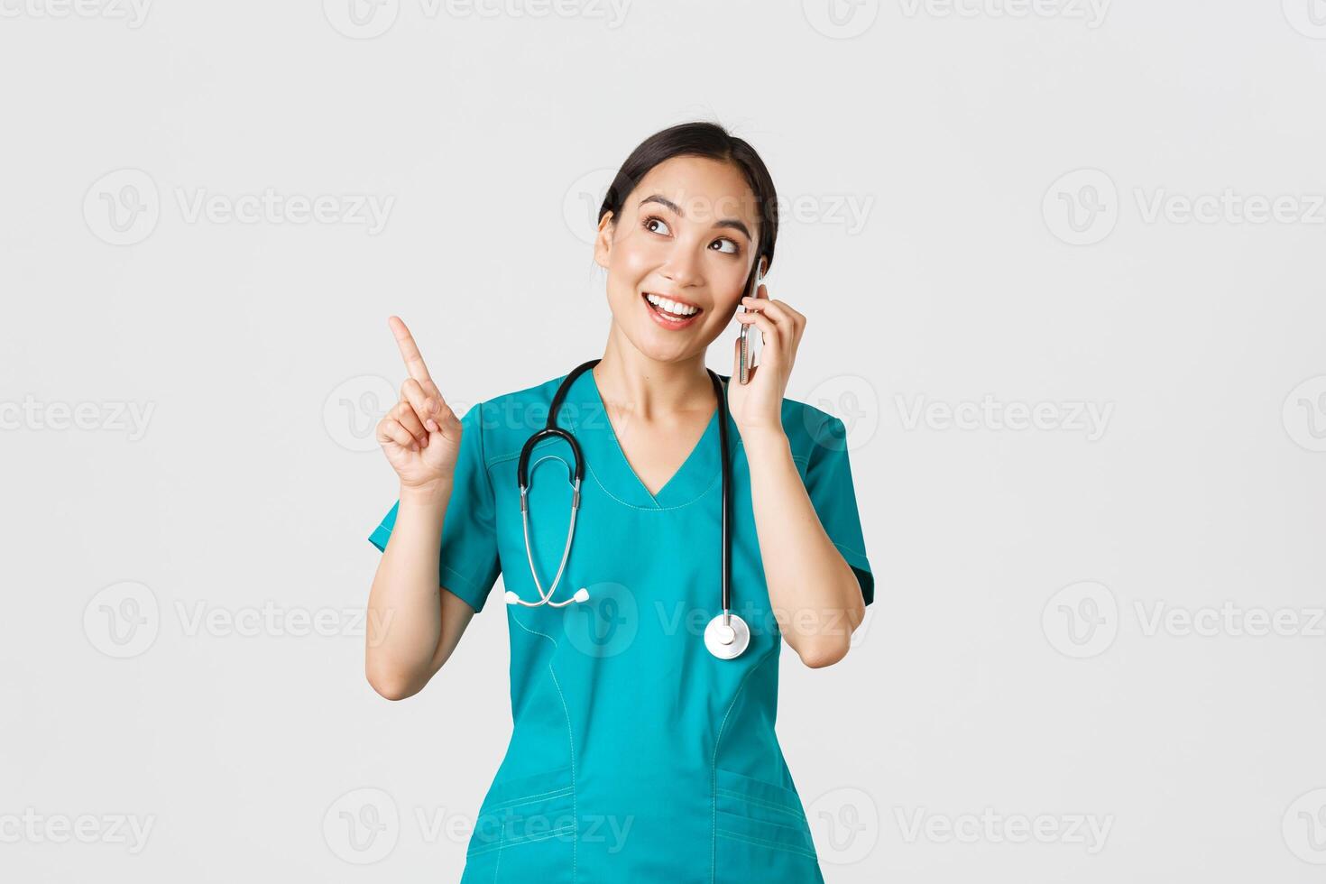 Covid-19, healthcare workers and preventing virus concept. Portrait of young pretty asian female doctor, nurse in scrubs making online order, talking on phone and pointing upper left corner, smiling photo