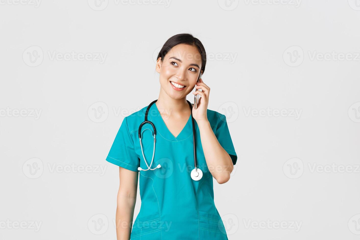 Covid-19, healthcare workers and preventing virus concept. Pretty smiling asian female doctor, physician in scrubs having conversation, talking on phone and looking upper left corner dreamy photo