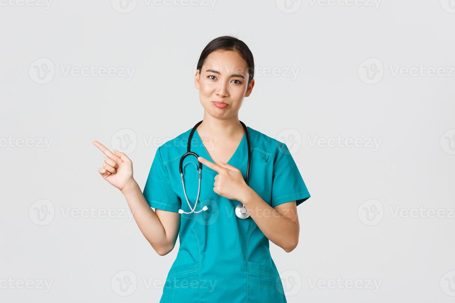 Covid-19, healthcare workers, pandemic concept. Skeptical and doubtful asian female nurse in scrubs pointing upper left corner and grimacing unamused, dont like product, being hesitant photo