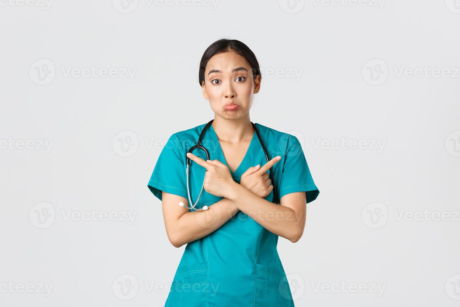 Covid-19, healthcare workers, pandemic concept. Indecisive and clueless asian female nurse, intern dont know what choose, pointing fingers sideways and shrugging unaware, white background photo