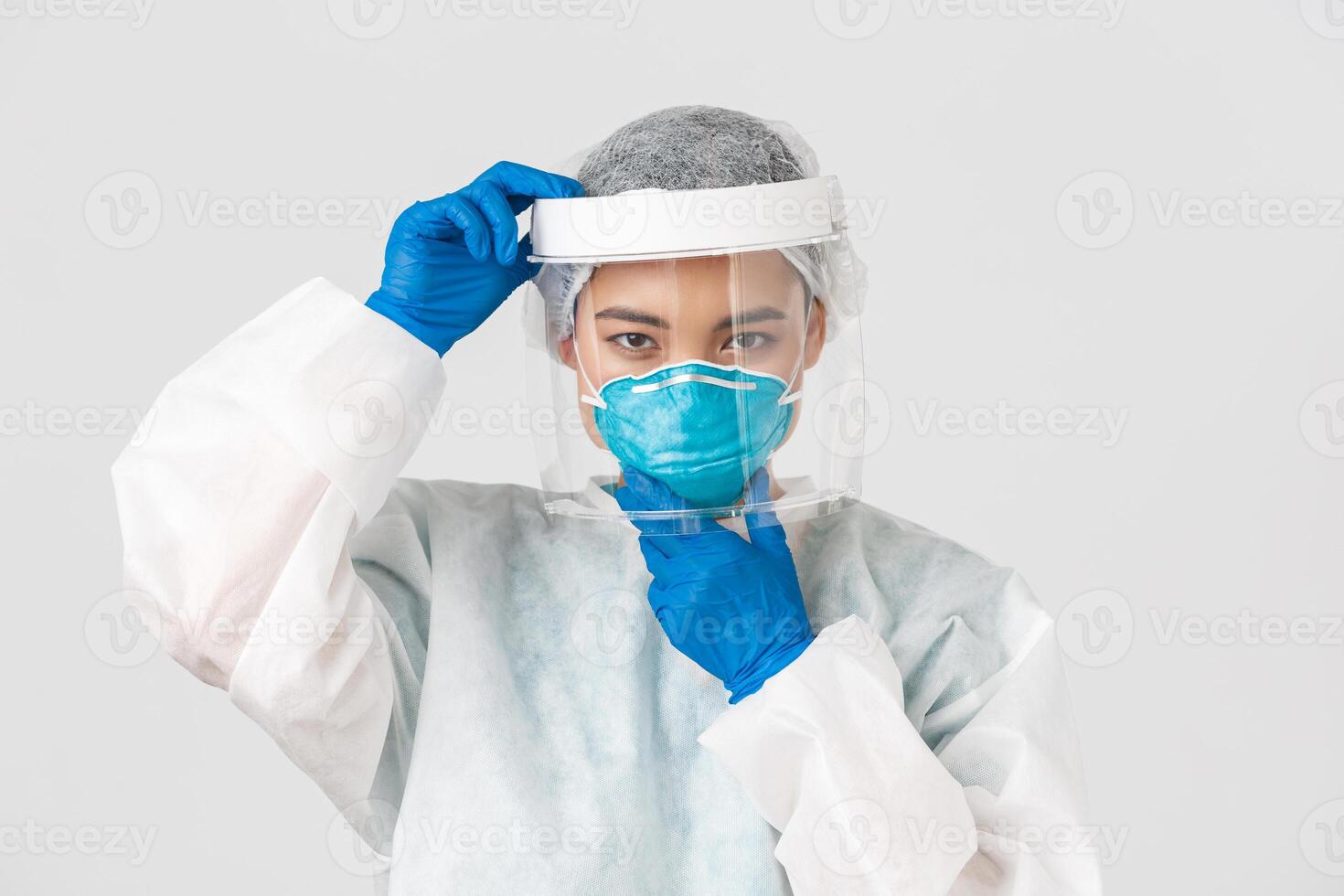 Covid-19, coronavirus disease, healthcare workers concept. Confident serious-looking female asian doctor, put on face shield and respirator, personal protective equipment, white background photo