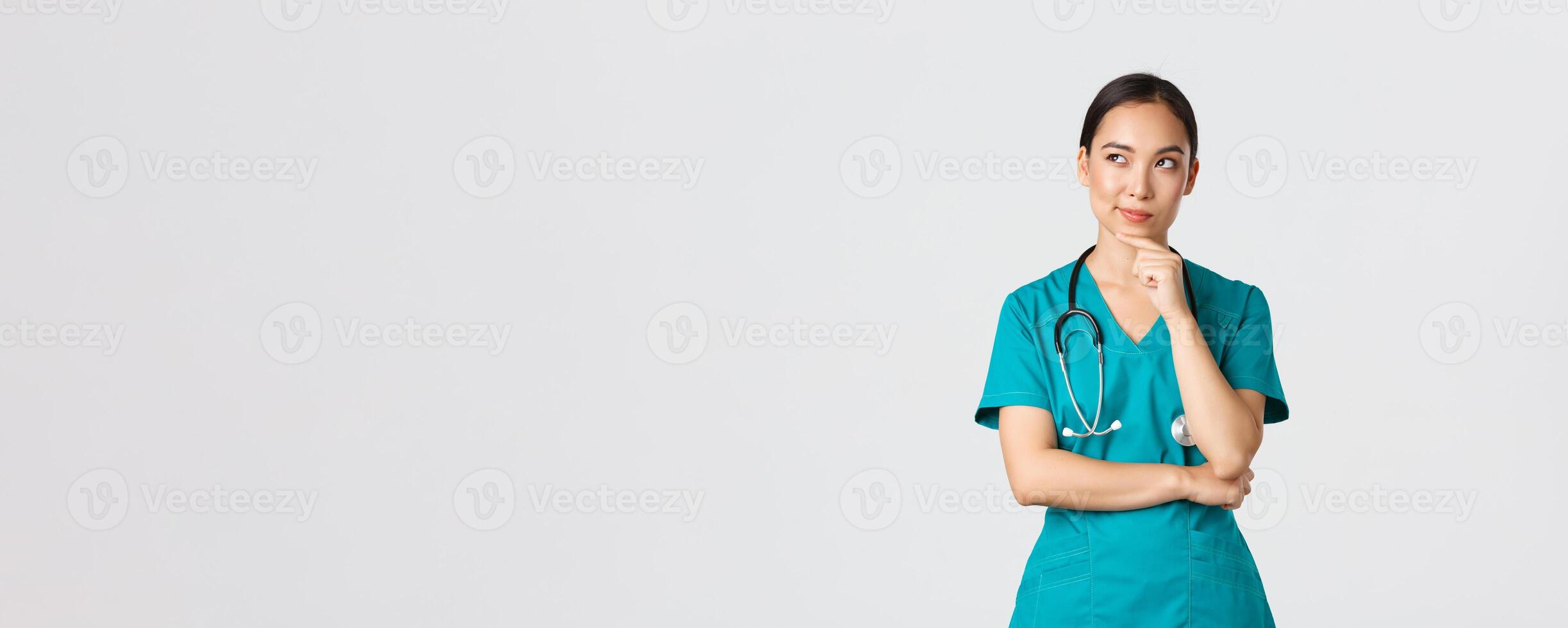 Covid-19, healthcare workers, pandemic concept. Thoughtful smart asian nurse in scrubs looking away and thinking, smiling pleased. Doctor have interesting idea, pondering over white background photo