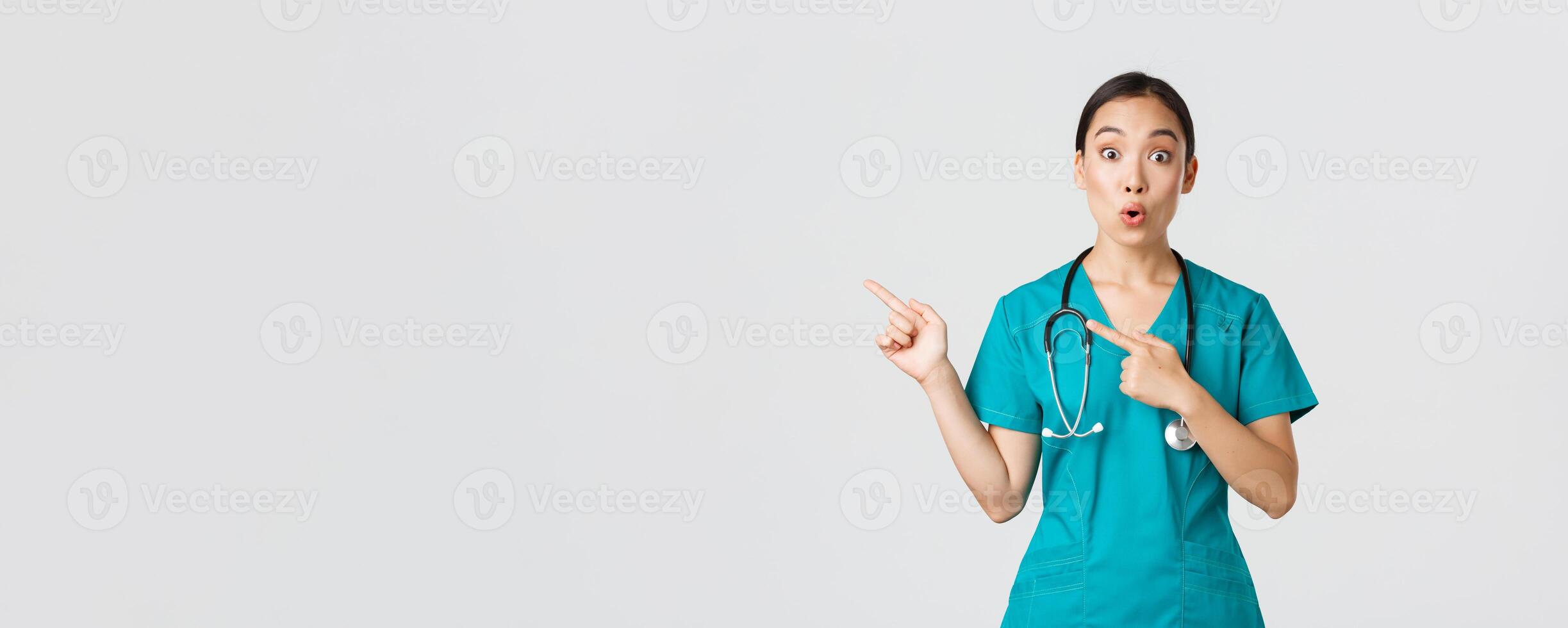 Covid-19, healthcare workers, pandemic concept. Impressed and amazed female asian nurse in scrubs asking question with intrigued expression. Doctor looking thrilled and pointing upper left corner photo