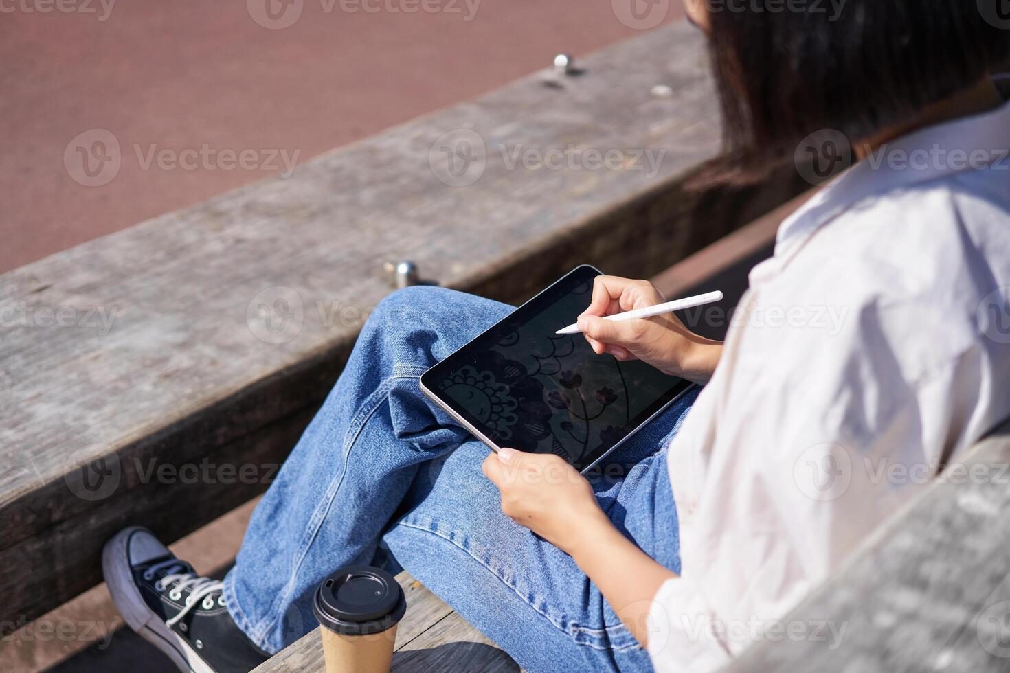 Cropped shot of female hands drawing on digital graphic tablet with pen, sitting with cup of coffee outdoors on bench photo