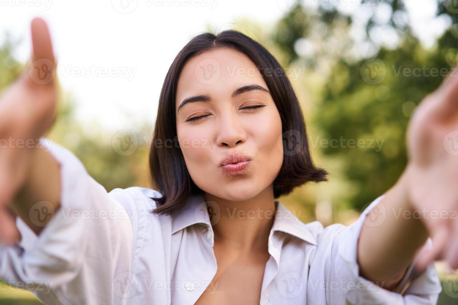 People and mobile connection. Happy young asian woman takes selfie on smartphone, holds camera with hands, poses in park on summer day photo