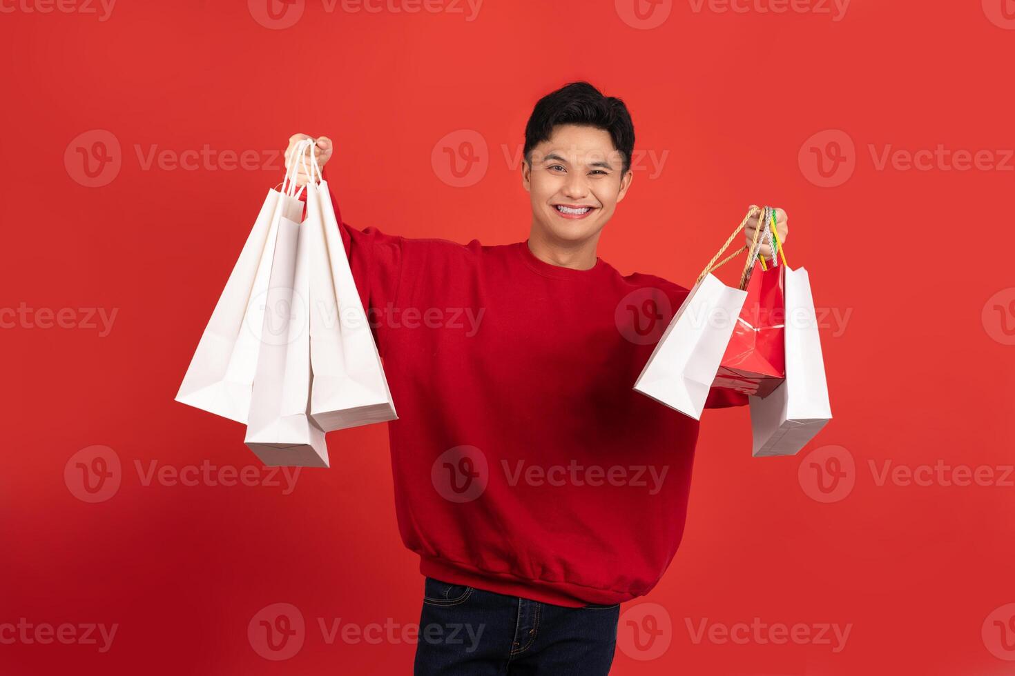 Cheerful young Asian man carrying many shopping bags on red background. photo