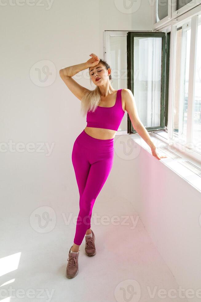mujer en vibrante rosado Deportes atuendo en pie siguiente a ventana con palma en frente foto