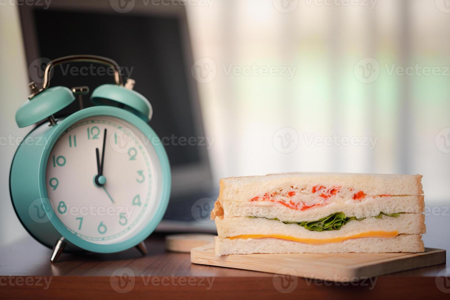 Sandwich and alarm clock displaying noon time on wooden table in office photo