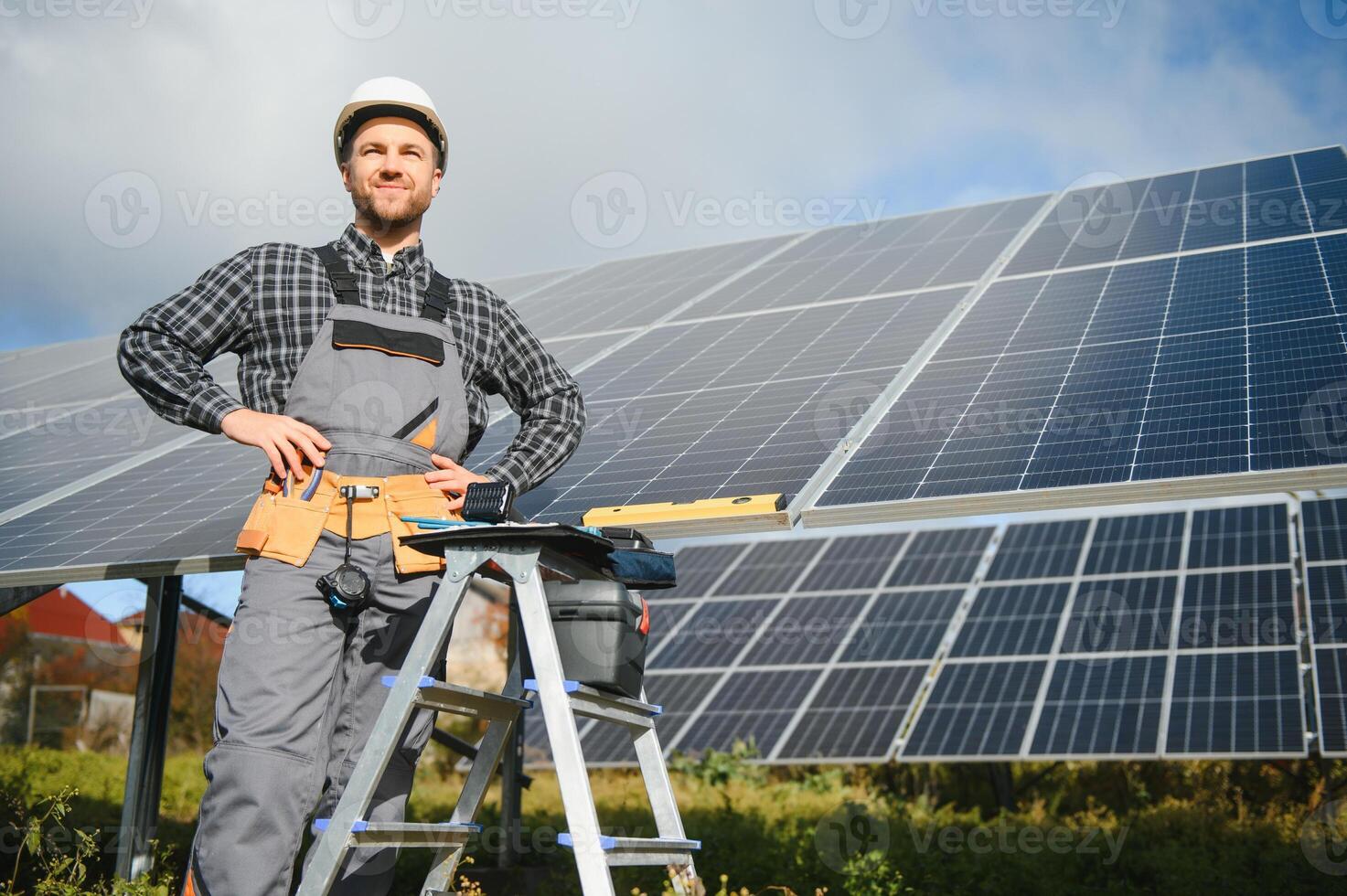 Portrait of smiling confident engineer technician with electrical screwdriver, standing in front of unfinished high exterior solar panel photo voltaic system