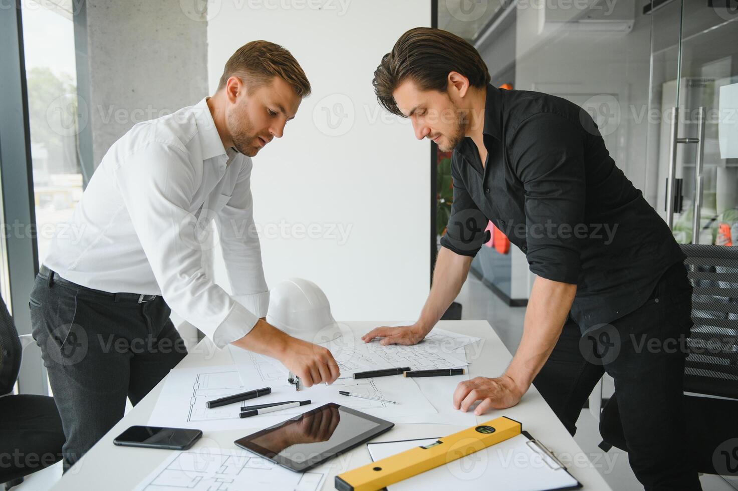two building designers standing in a modern office leaning over a desk discussing blueprints together photo