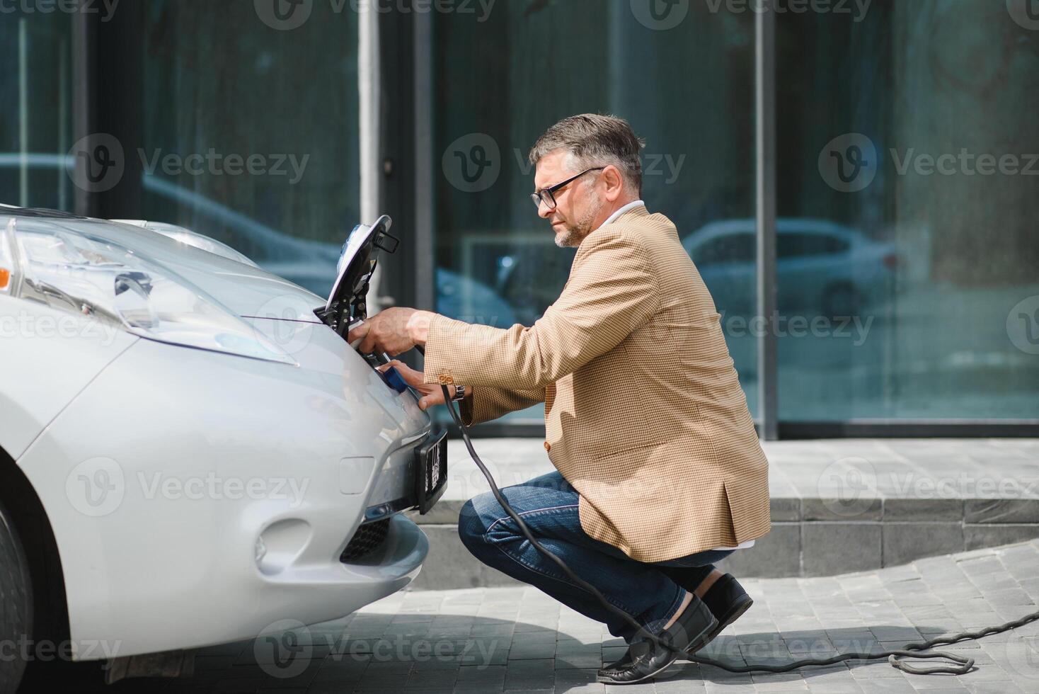 Hansome bearded man sitting near his new modern electric car and holding plug of the charger, while car is charging at the charging station photo