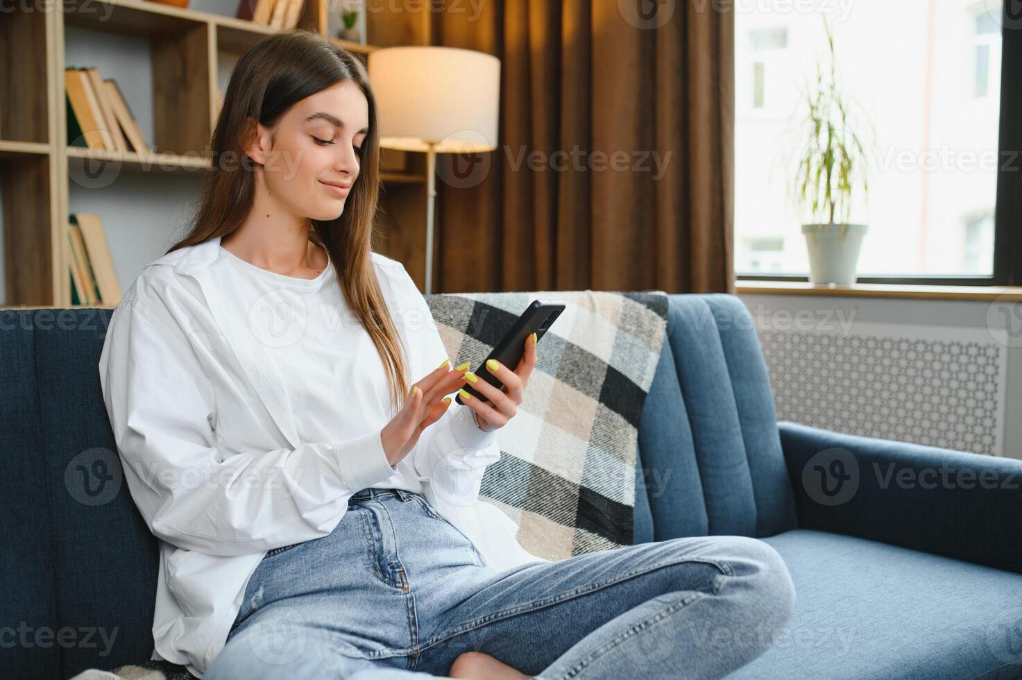 Young happy caucasian woman she 20s in casual clothes hold in hand use mobile cell phone sit on blue sofa indoor rest at home in own room apartment. People lifestyle concept photo