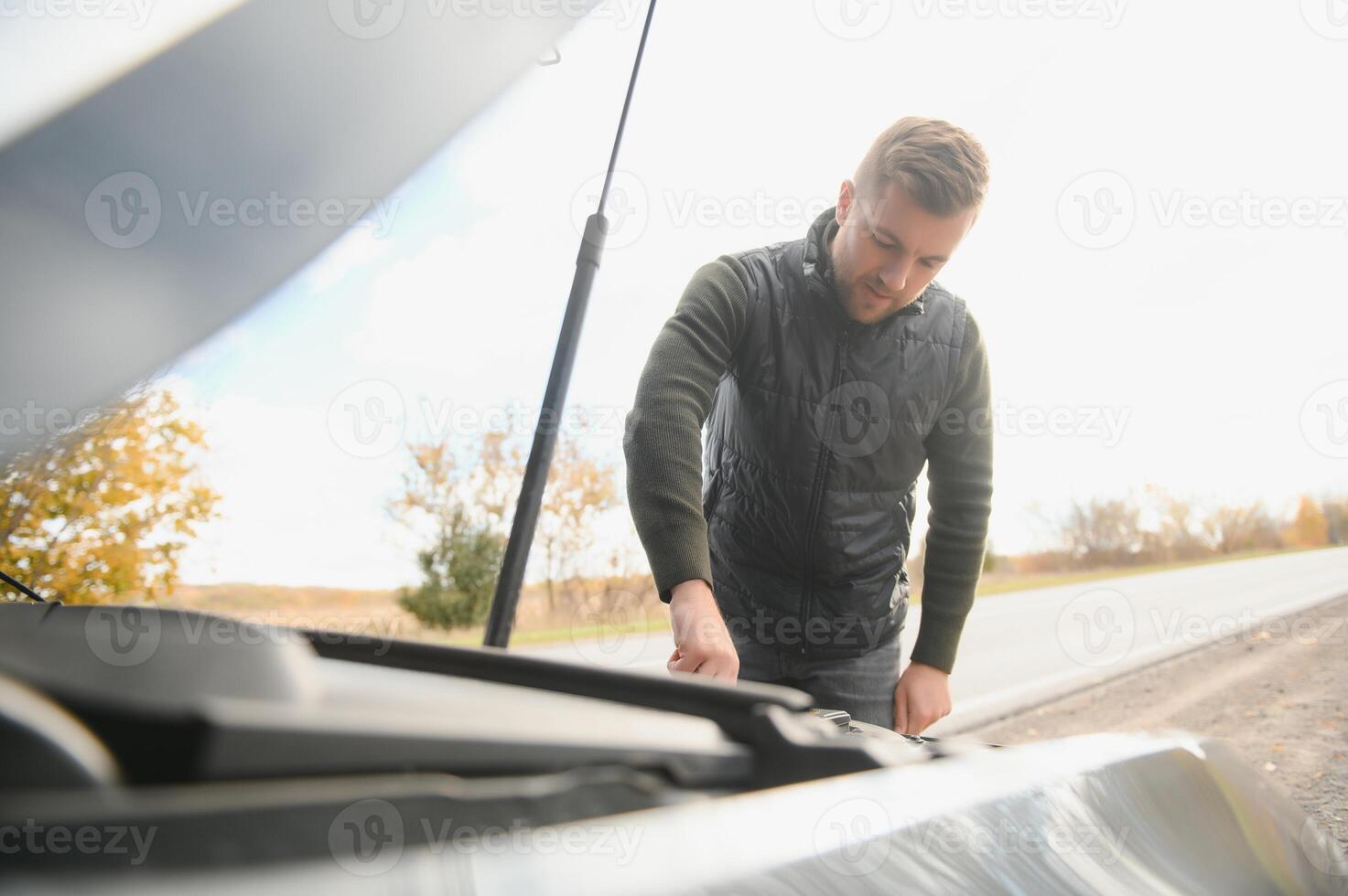 Man repairing a broken car by the road. Man having trouble with his broken car on the highway roadside. Man looking under the car hood. Car breaks down on the autobahn. Roadside assistance concept. photo