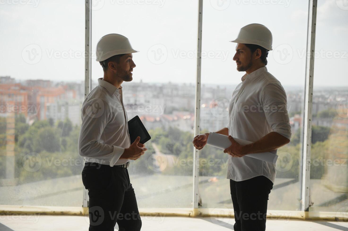 A front view of two smart architects with white helmets reviewing blueprints at a construction site on a bright sunny day photo