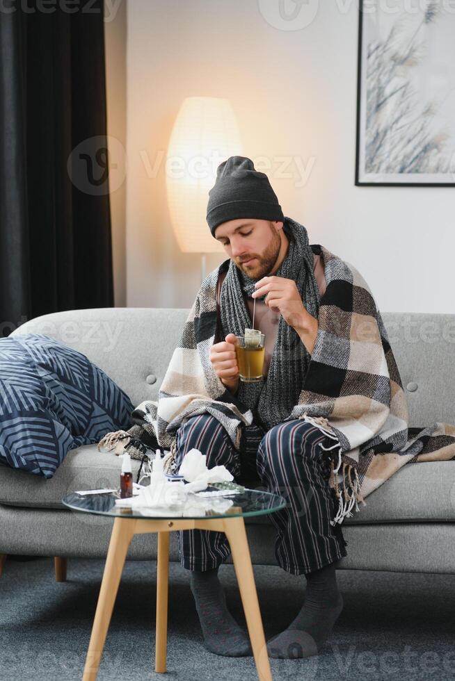 It's cold at home in wintertime. Man freezing in his house in winter because of broken thermostat. young guy wrapped in woolen plaid shivering while sitting on sofa in living room interior photo