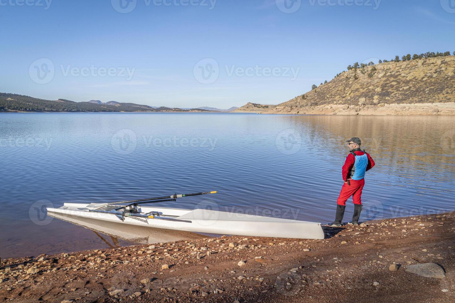 mayor masculino remero y un costero remo cáscara es aterrizaje en un rocoso apuntalar de carretero lago en otoño o invierno paisaje en del Norte Colorado. foto