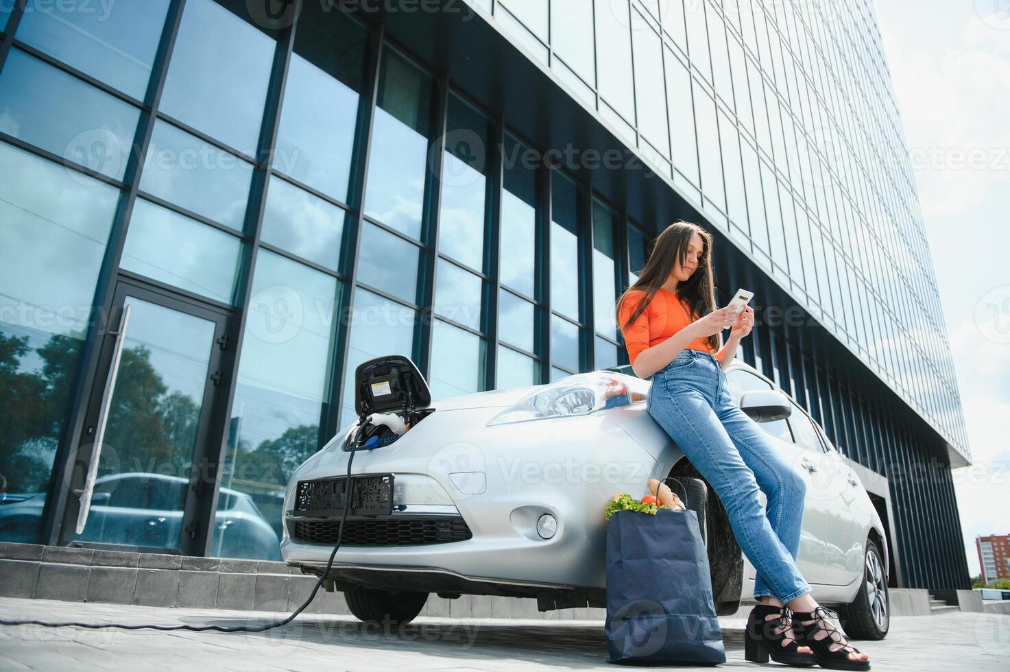 utilizando teléfono inteligente mientras espera. mujer en el eléctrico carros cargar estación a tiempo de día. marca nuevo vehículo. foto