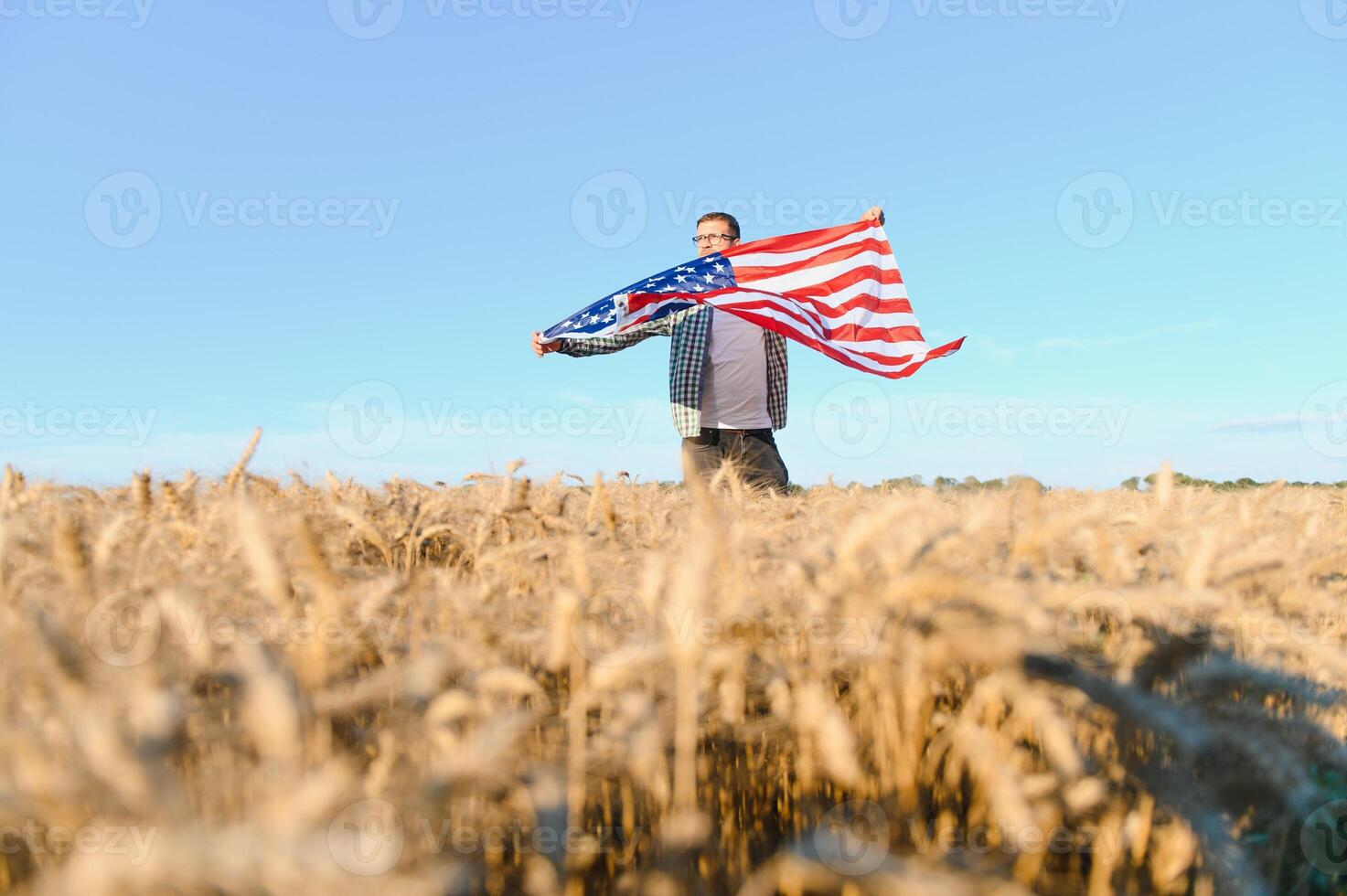 Young patriotic farmer stands among new harvest. Boy walking with the american flag on the wheat field celebrating national independence day. 4th of July concept. photo