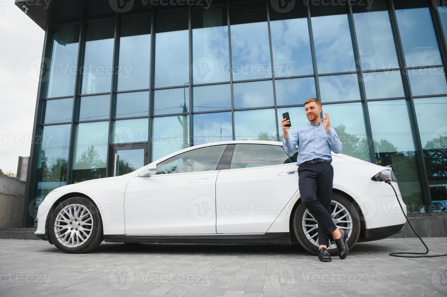 hermoso hombre en negocio traje surf Internet en moderno teléfono inteligente mientras esperando eléctrico coche a cargar. foto