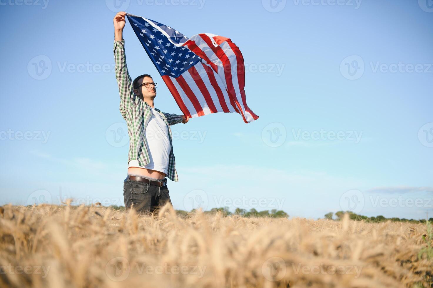 Young patriotic farmer stands among new harvest. Boy walking with the american flag on the wheat field celebrating national independence day. 4th of July concept. photo