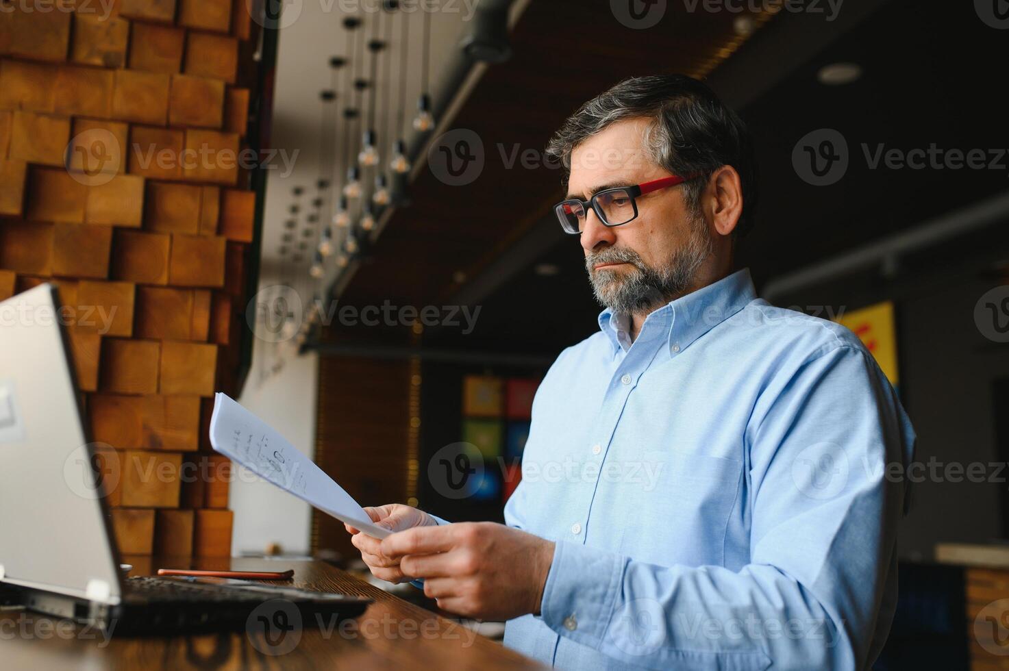 Positive senior bearded man with grey hair drinking coffee and using laptop at cafe, copy space. Stylish aged businessman in burgundy jacket enjoying his tea while working online. photo