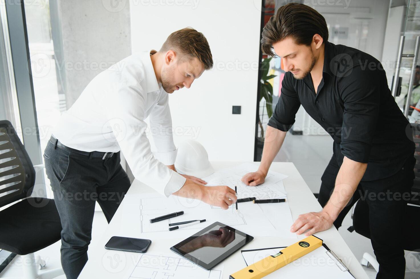 two building designers standing in a modern office leaning over a desk discussing blueprints together photo