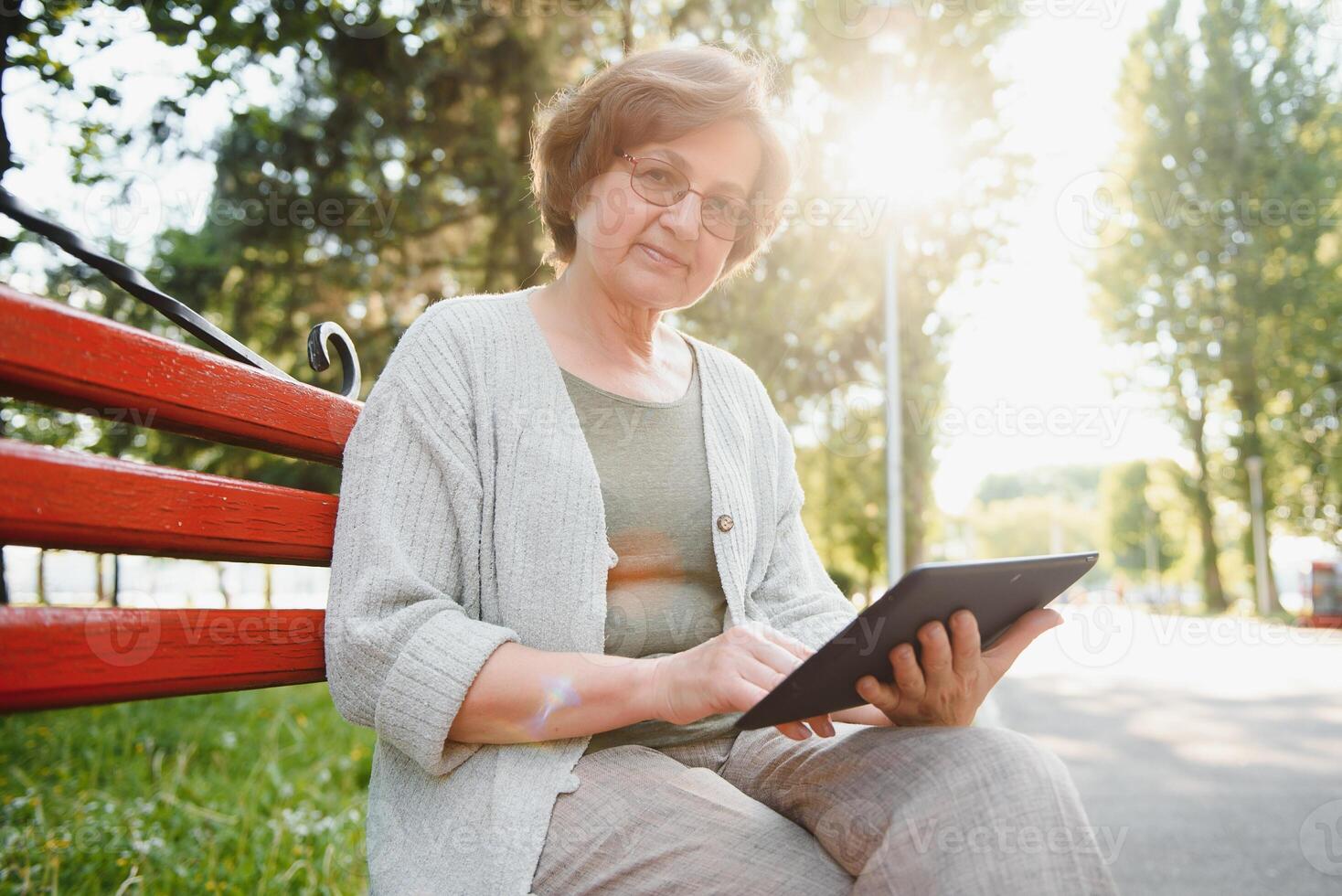 Positive confident old lady posing with tablet in park. Senior grey haired woman in casual sitting on park bench and using tablet. Wireless connection concept photo