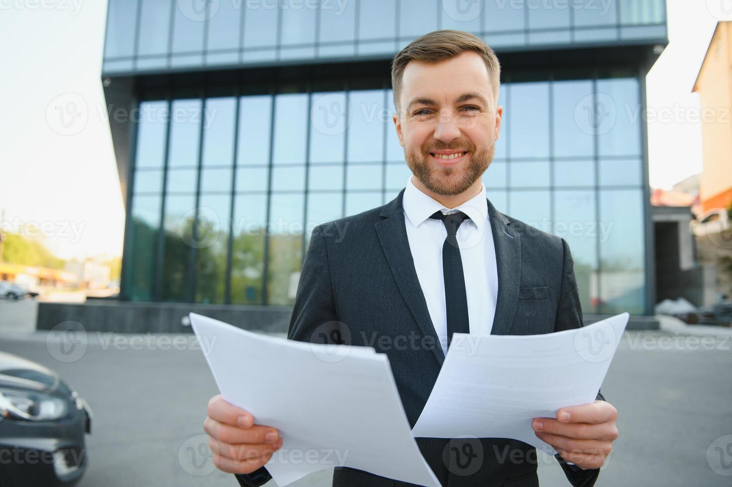 Portrait of handsome modern male businessman holding folder with documents on background of urban buildings and offices. photo