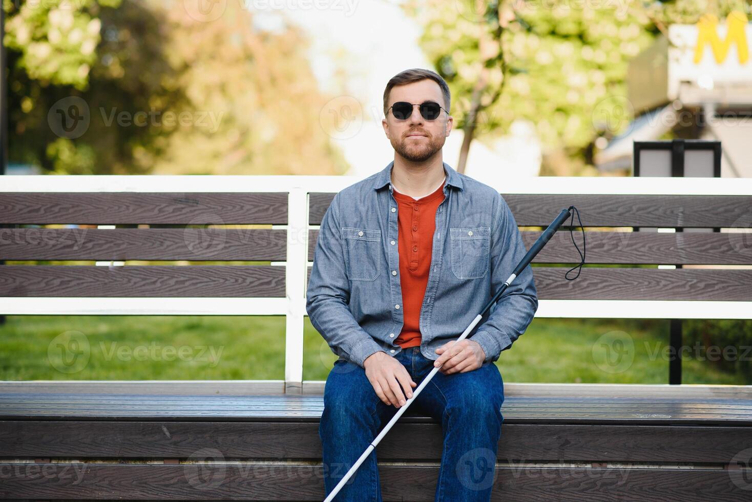 Visually impaired man with walking stick, sitting on bench in city park. Copy space photo