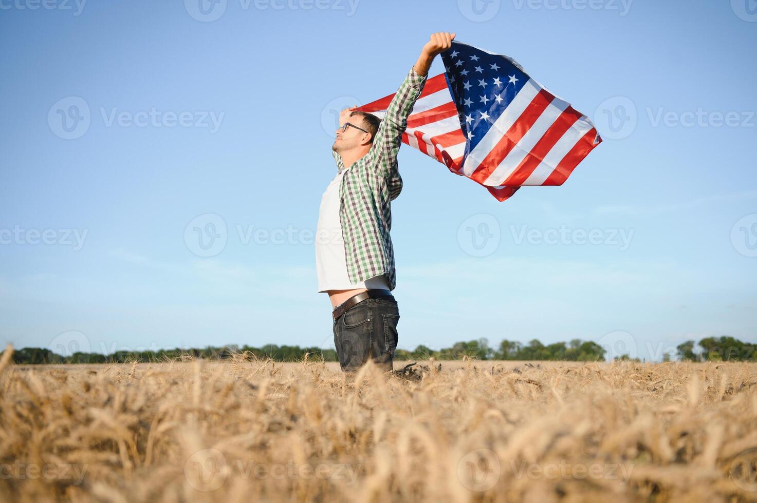 Young patriotic farmer stands among new harvest. Boy walking with the american flag on the wheat field celebrating national independence day. 4th of July concept. photo