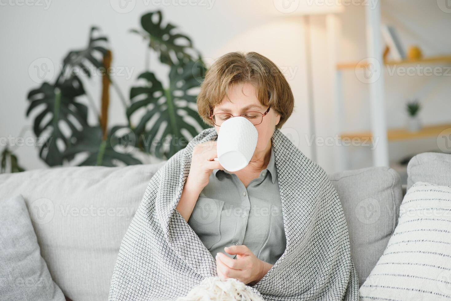 Smiling mature elder 65s woman sitting relaxing with cup of tea, coffee. Senior mid age stylish look woman with eyeglasses portrait with cup looking away at modern home photo