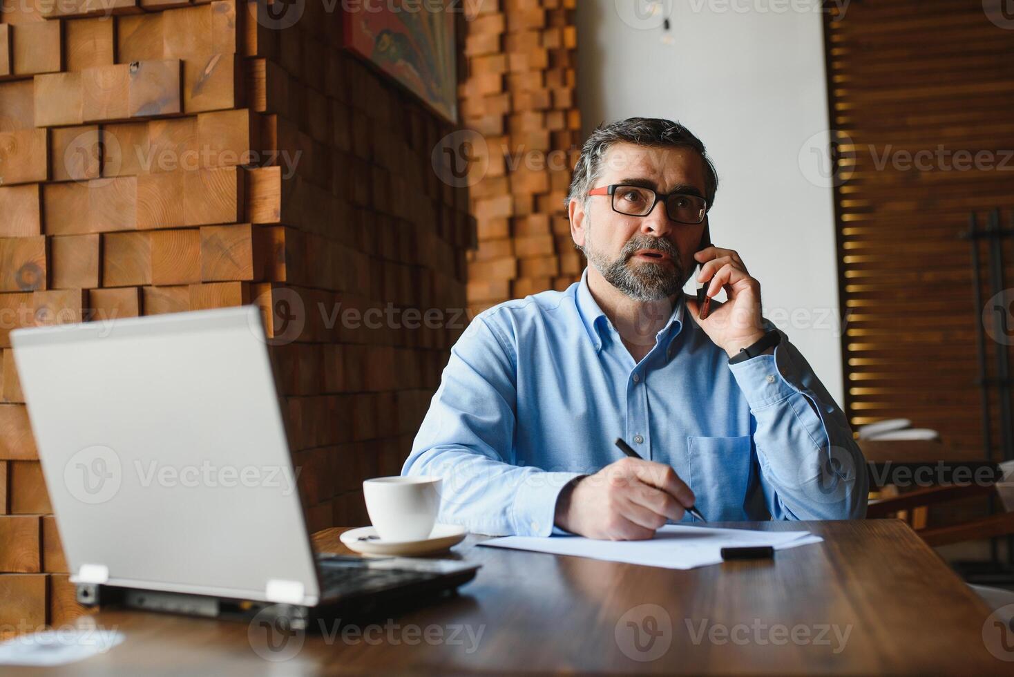 Positive senior bearded man with grey hair drinking coffee and using laptop at cafe, copy space. Stylish aged businessman in burgundy jacket enjoying his tea while working online. photo