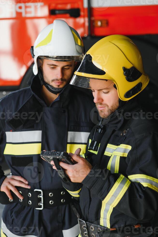 Two professional firefighters with uniforms and protective helmets posing infront of a firetruck. photo