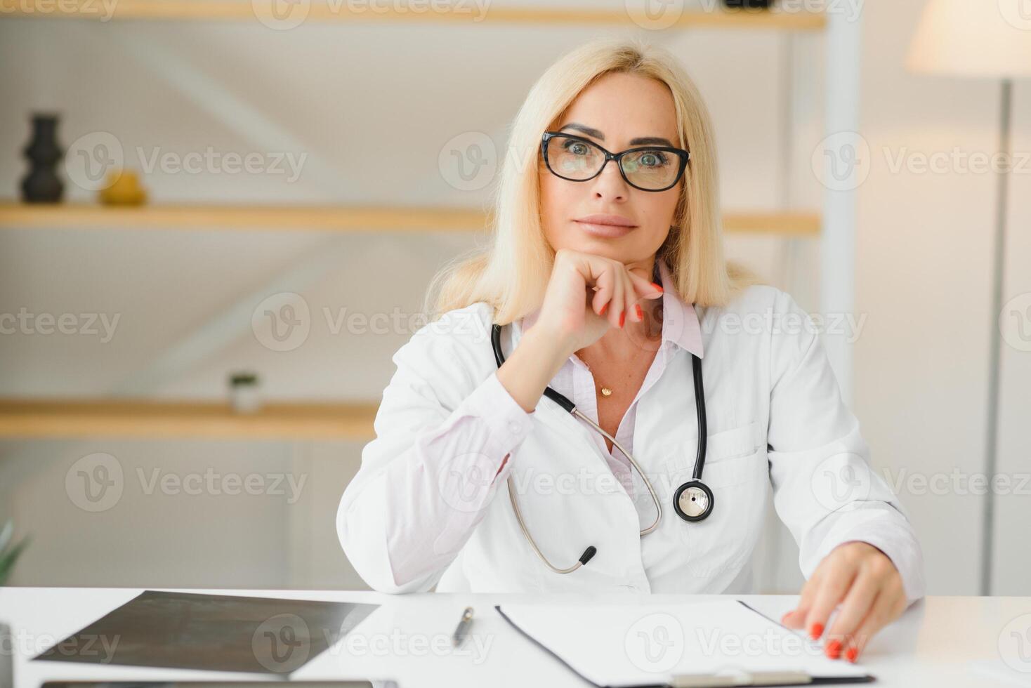 Portrait of middle age female doctor is wearing a white doctor's coat with a stethoscope around her neck. Smiling physician standing at private clinic photo