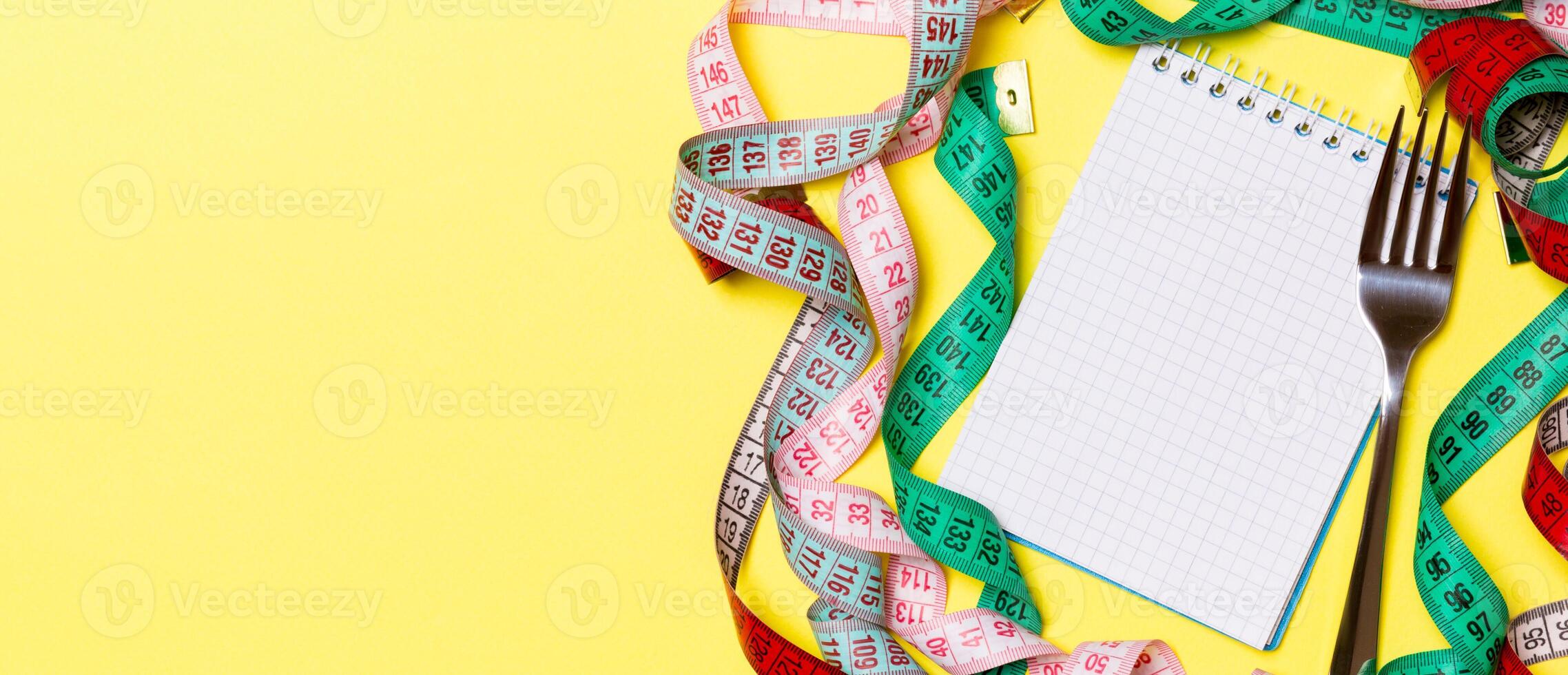 Fitness concept with empty space for your design on yellow background. Top view of notepad and fork surrounded with colorful measure tapes photo